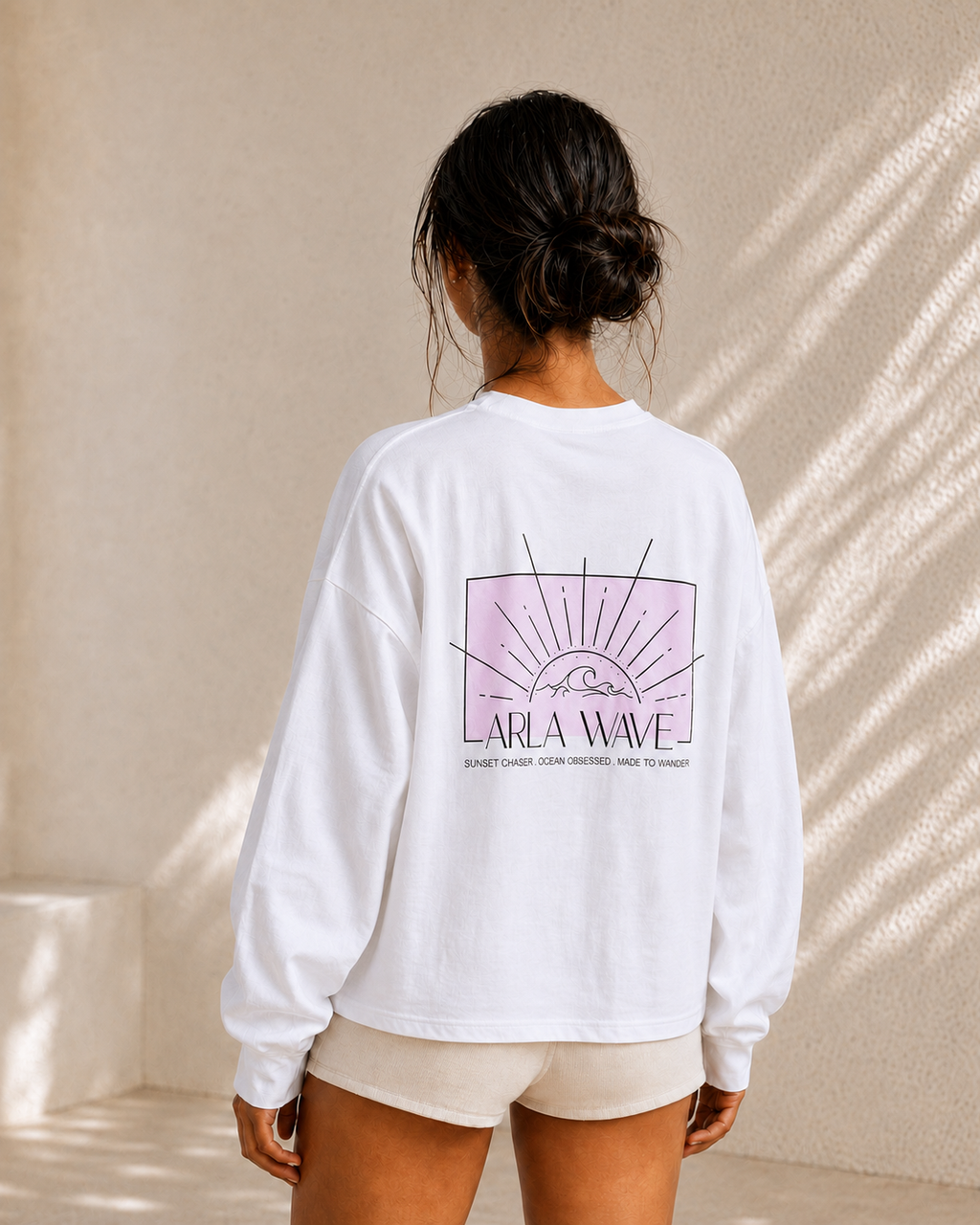 A woman viewed from behind wearing a white long sleeve box-cut graphic sweatshirt with a pink and black 'Arla Wave' sunburst design on the back, paired with light beige shorts, standing against a textured beige wall with soft diagonal shadows.